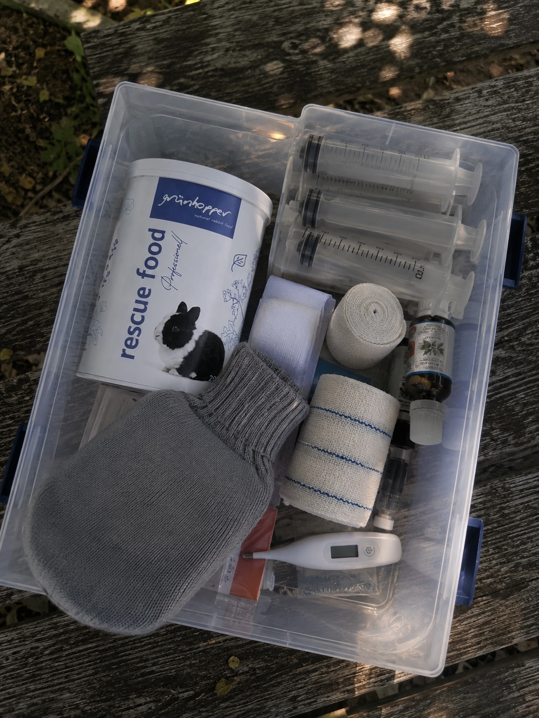 Geöffnete Kunststoffbox mit Hausapotheke-Inhalt auf einem Gartentisch: Päppelbrei-Pulver, Spritzen, Mullbinde, Thermometer und kleine Wärmflasche